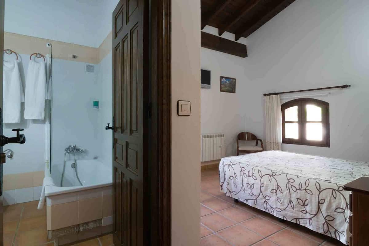 A bedroom with a bed, a window, and a bathroom with a bathtub and towel rack.