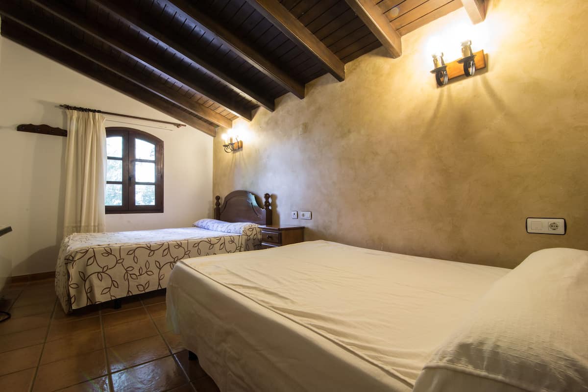 A bedroom with two beds, wooden beams on the ceiling, a window with curtains, and a wall-mounted shelf with lights.