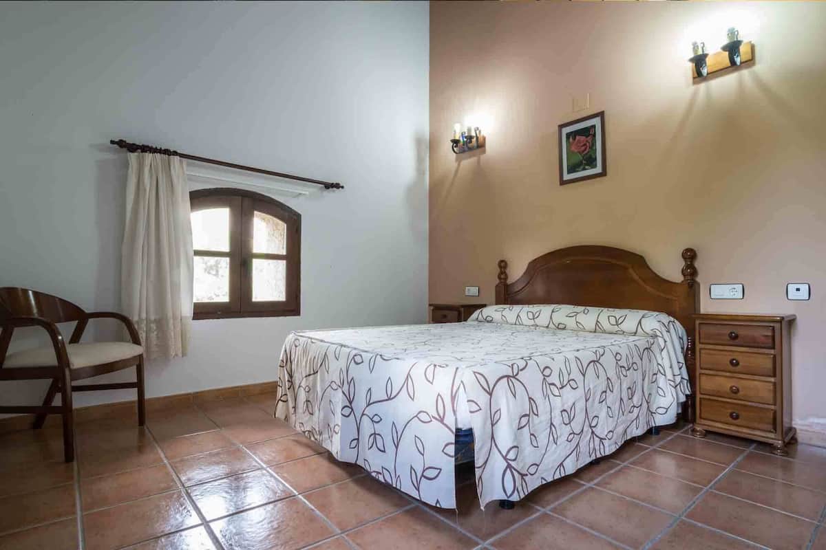 A bedroom with a bed, a chair, a nightstand, a dresser, a window with curtains, and a wall-mounted light fixture.