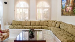 Uma sala de estar com um amplo sofá bege, uma poltrona pequena e uma mesa de centro com um vaso de flores. Há duas janelas com cortinas leves e um quadro na parede.