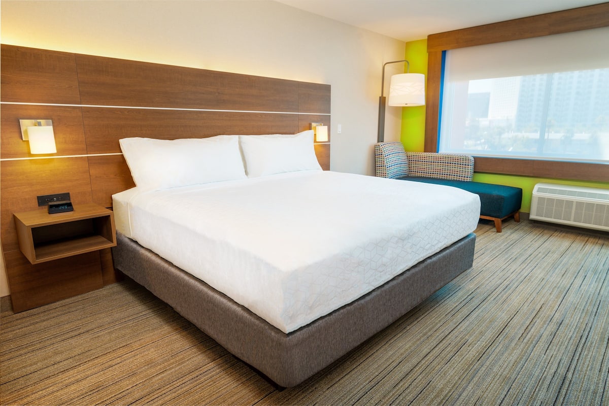 Une chambre d’hôtel avec un grand lit, des tables de chevet, une petite table, une chaise, une fenêtre avec vue et une lampe fixée au mur.