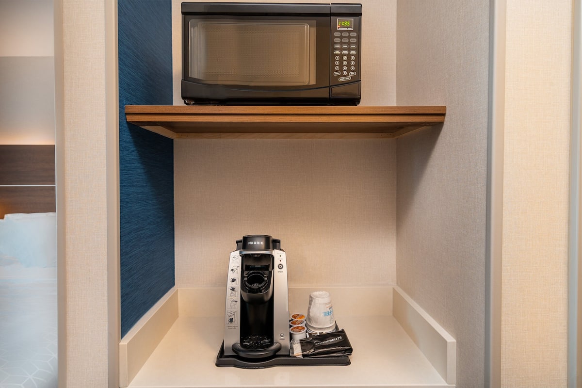 Une petite cuisine équipée d’un four à micro-ondes posé sur une étagère, d’une machine à café Keurig et d’une bouteille d’eau.
