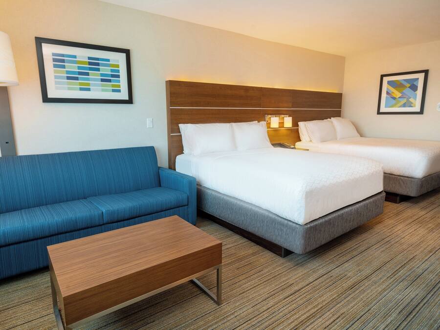 Quarto de hotel com sofá azul, mesa de centro de madeira, cama com roupa de cama branca e duas mesas de cabeceira com luminárias.