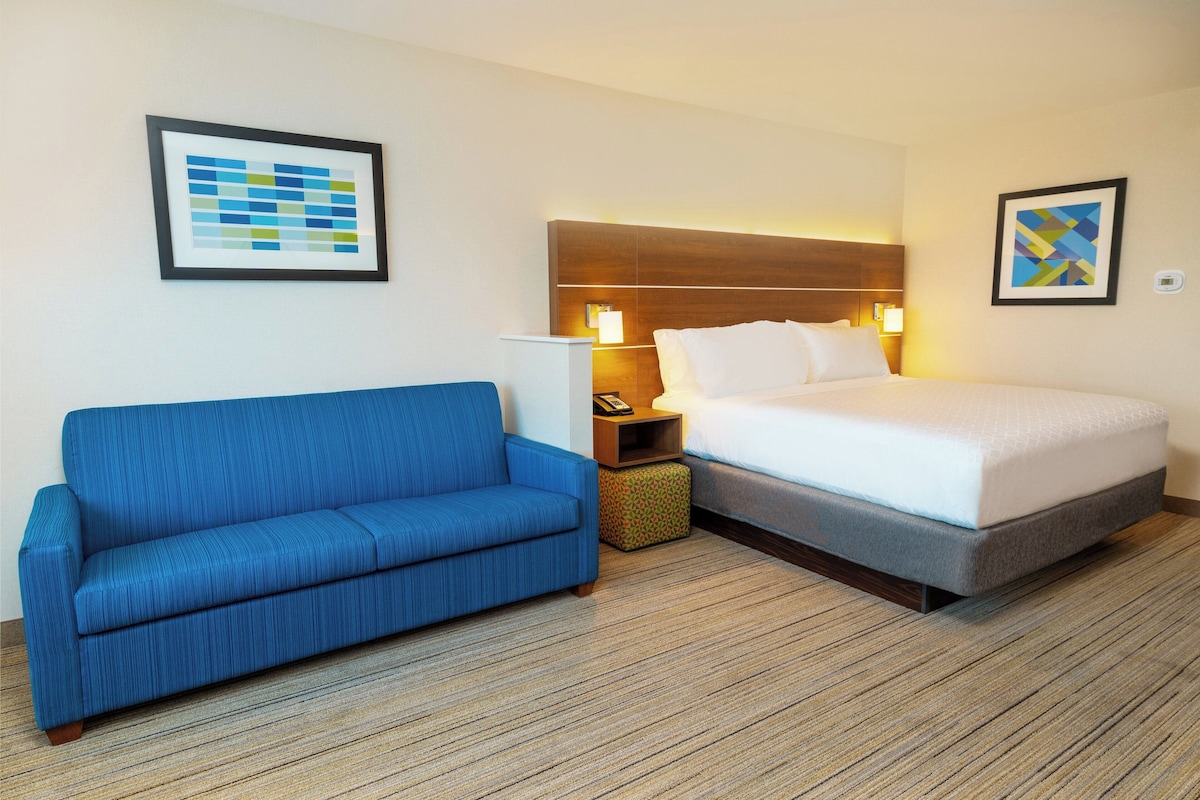 Een hotelkamer met een bed, een blauwe bank, een nachtkastje met een lamp, en twee ingelijste foto's aan de muur.