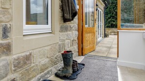 A stone building with a wooden door and windows, a pair of boots on a mat, and a coat hanging on the wall. - 3 bedroom accommodation in Hathersage (Hathersage)