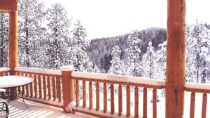 A wooden deck with a table and chairs, surrounded by snow-covered trees and a forested hill.