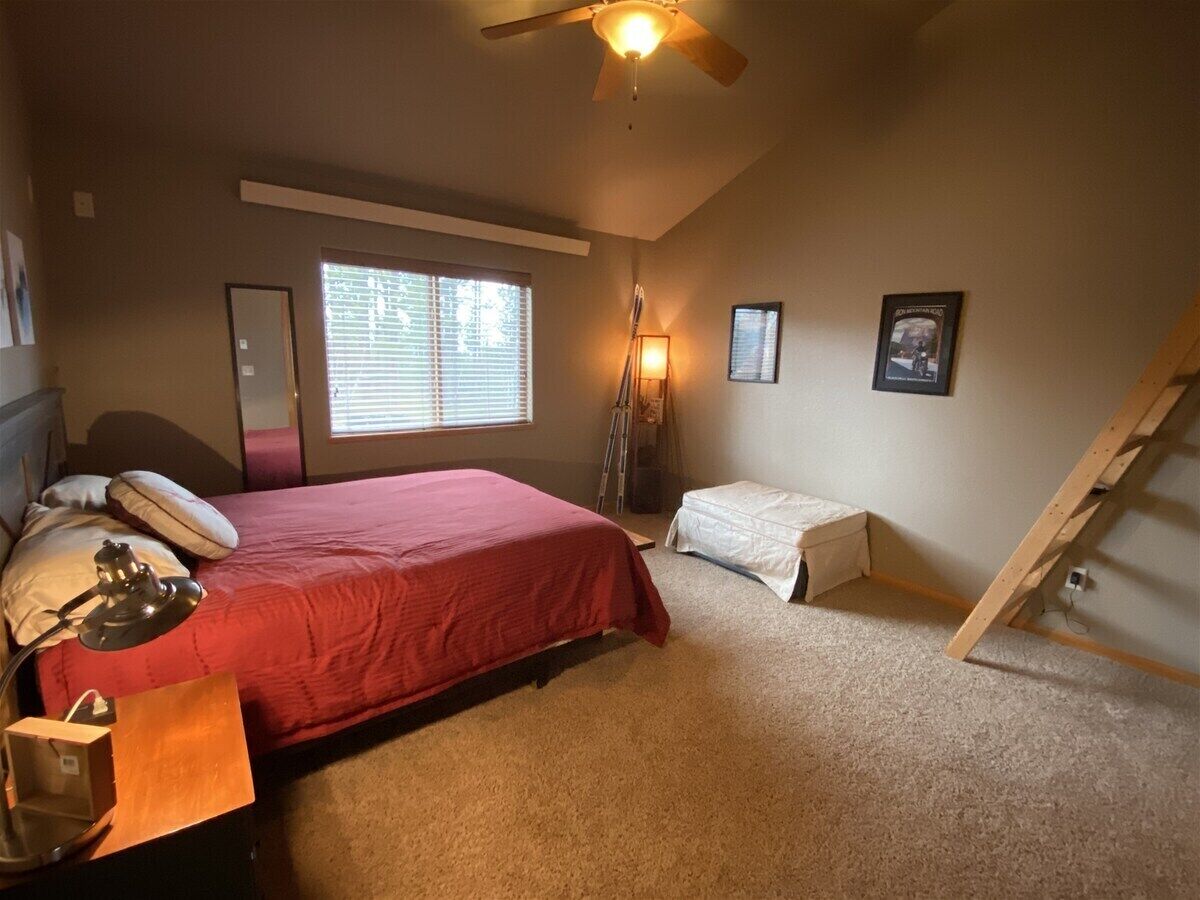 A bedroom with a bed, a desk, a lamp, a ladder, a window with blinds, and a ceiling fan.