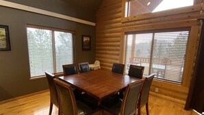A log cabin interior with a wooden dining table surrounded by chairs, large windows with blinds, and a framed picture on the wall.