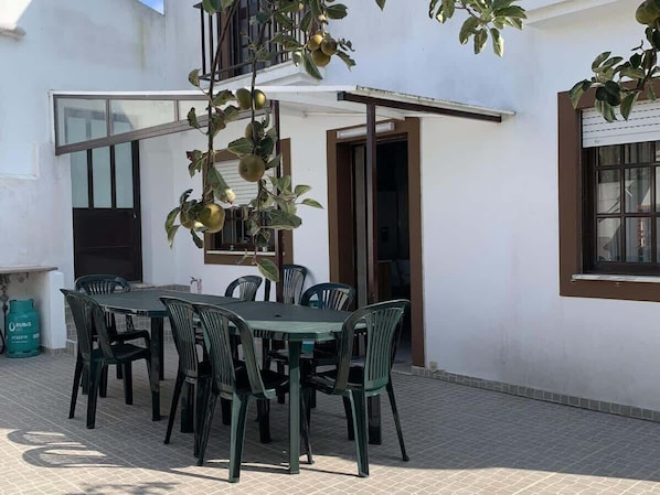 Une terrasse avec une table et des chaises, une plante suspendue et un bâtiment blanc doté d’une fenêtre et d’une porte.