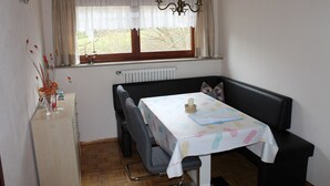 Un coin repas avec une table recouverte d’une nappe à motifs floraux, un banc noir rembourré et un radiateur placé sous une fenêtre ornée de rideaux.