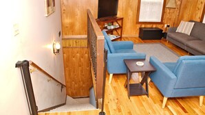 A wooden-floored living room with a blue sofa, a television on a wooden stand, and a small table with a lamp. - Morehead Cabin on the lake (Morehead)