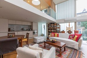 A modern living room with a sofa, coffee table, and chairs. There is a kitchen area with cabinets and appliances. A bookshelf and a lamp are also visible.