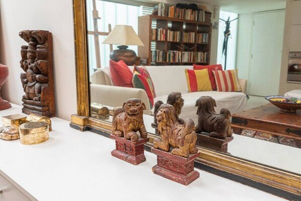 A room with a white sofa, a wooden sculpture, and a mirror reflecting a bookshelf and a lamp.