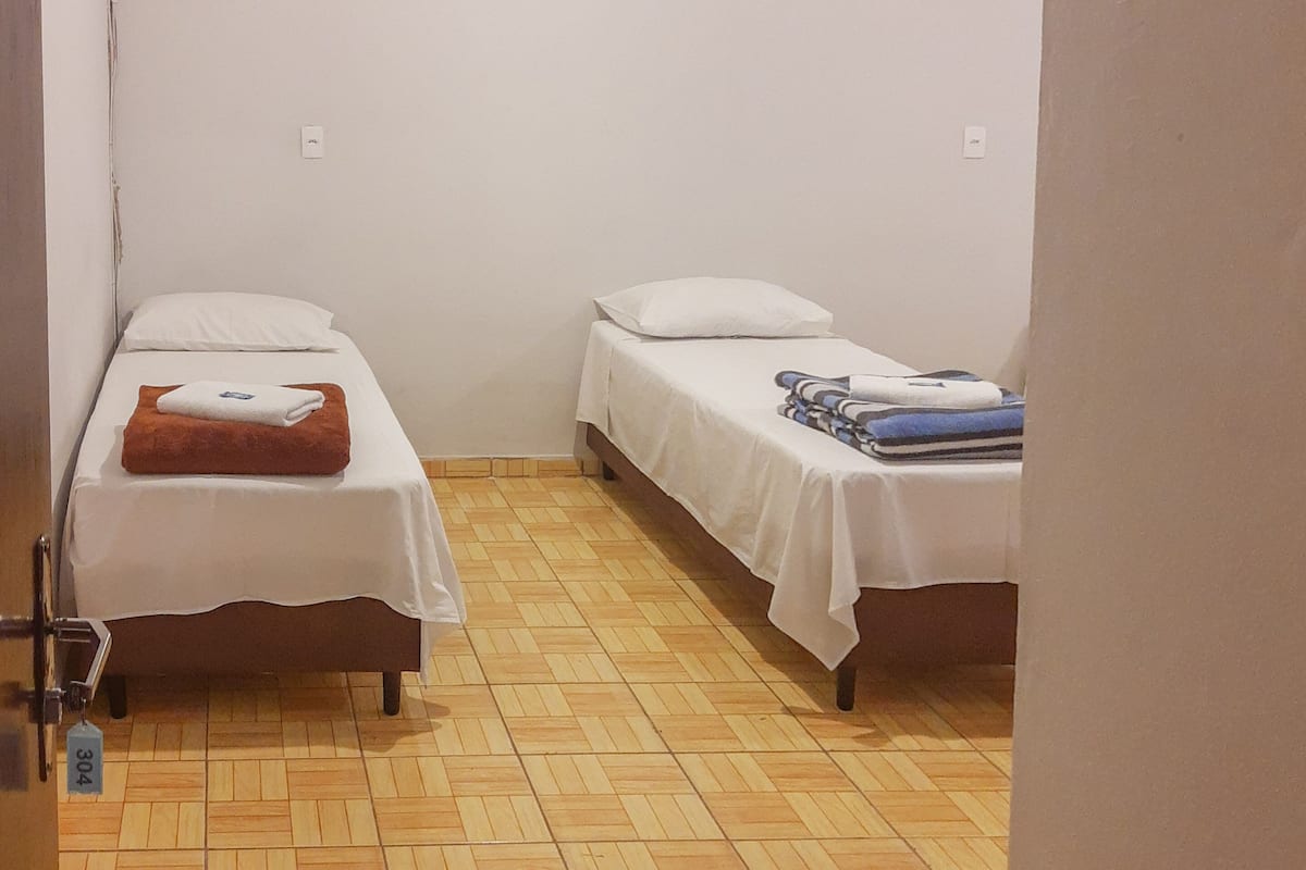 Dos camas individuales con sábanas blancas, piso de madera y una puerta con cerradura.