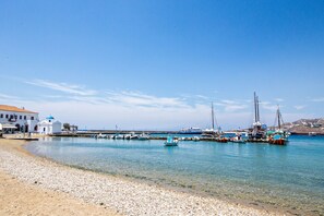 A coastal scene with a pebbled beach, a white building with a blue dome, and several boats docked at a pier.