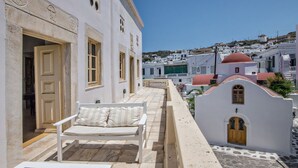 Un bâtiment blanc avec un banc et une porte, une petite structure blanche surmontée d’un dôme rouge, et une vue sur une ville avec des bâtiments blancs et une colline en arrière-plan.