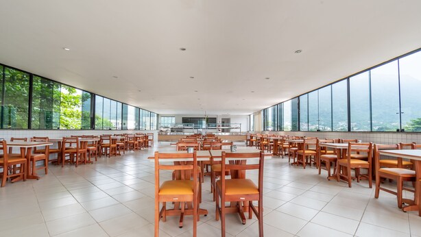 A spacious dining area with wooden tables and chairs, large windows offering a view of greenery and a cityscape. - OYO Residencial Muriqui Apart Hotel, Mangaratiba (Mangaratiba)