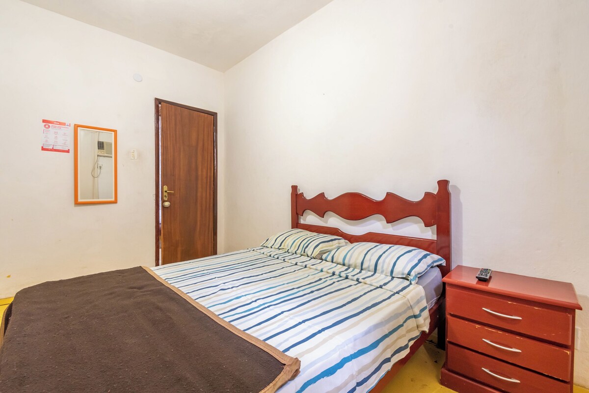 Um quarto com uma cama, cabeceira de madeira, mesa de cabeceira vermelha com gavetas e uma porta com espelho e um letreiro na parede.
