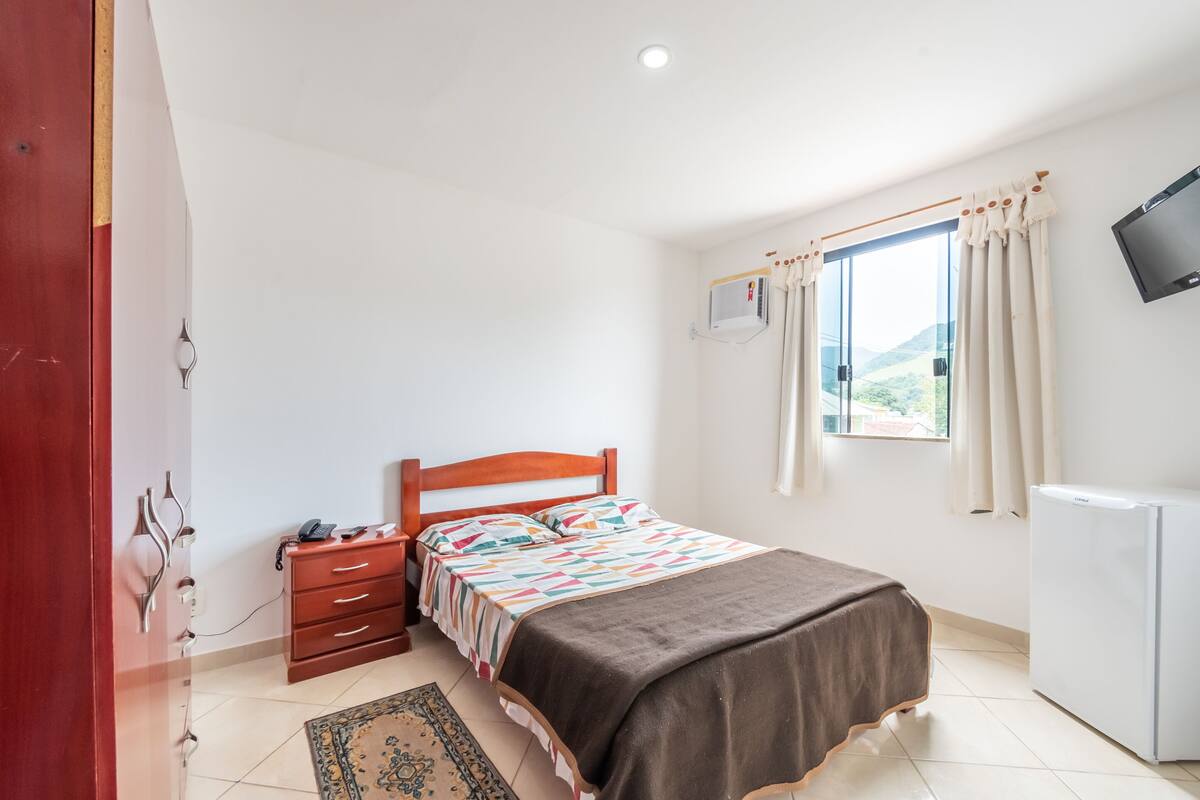 A bedroom with a bed, a wooden nightstand, a desk, a chair, a TV, and a window with curtains.
