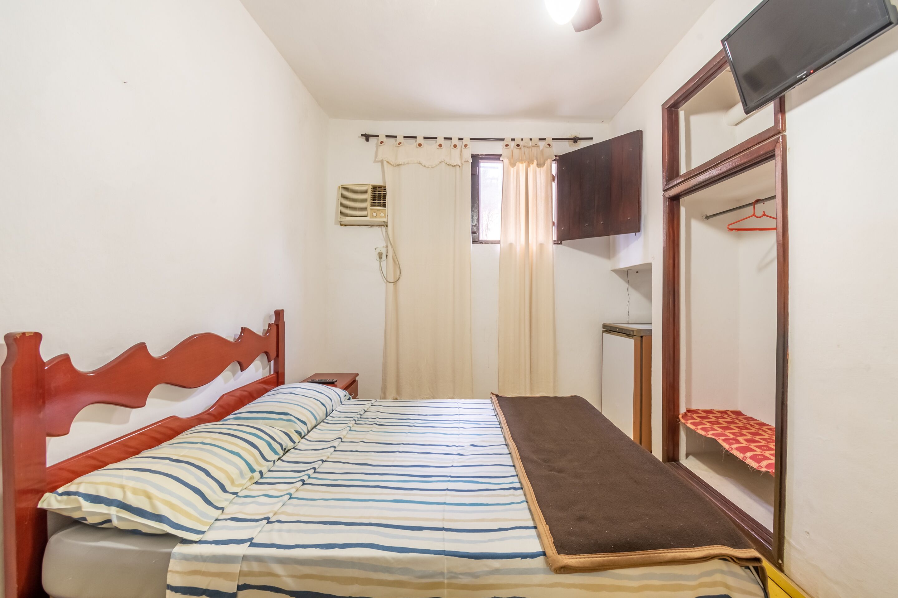 A bedroom with a bed, a wooden headboard, a bedside table, a TV mounted on the wall, and a window with curtains.