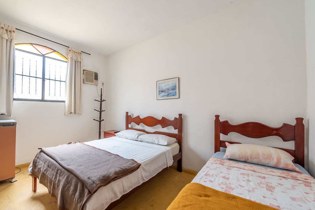 A bedroom with two beds, a window with curtains, a wall-mounted heater, and a picture.
