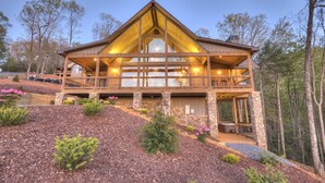 A two-story log cabin with a balcony, stone foundation, and a landscaped front yard.