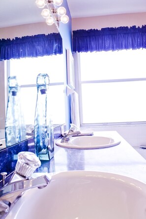 Ein Badezimmer mit einem großen Spiegel, zwei Glasflaschen, einem Waschbecken und einem Fenster mit blauem Vorhang.