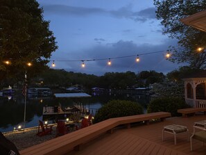 A wooden deck with outdoor seating, a view of a lake with boats, and string lights.