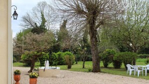 Une terrasse en gravier avec une table et des chaises blanches, entourée de différents arbres et arbustes.