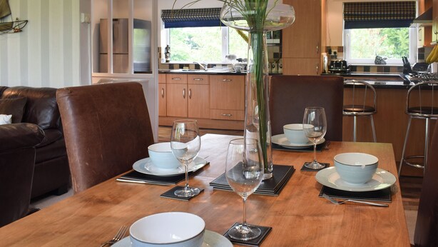 A dining area with a wooden table set for four, a vase with green plants, and a kitchen in the background.