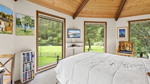 A bedroom with a large bed, wooden ceiling, and large windows offering views of a green landscape.