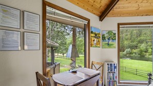 A room with a wooden table and chairs, a bookshelf, and large windows offering a view of a green landscape and a lake.