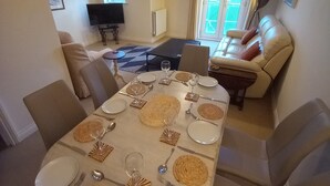 A dining area with a round table set for six, surrounded by beige chairs. There is a sofa with a cushion and a television in the background. - 47 Wetherby Way (Stratford-upon-Avon)