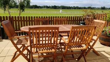 A wooden outdoor table and chairs set on a patio with a view of a green field and trees.