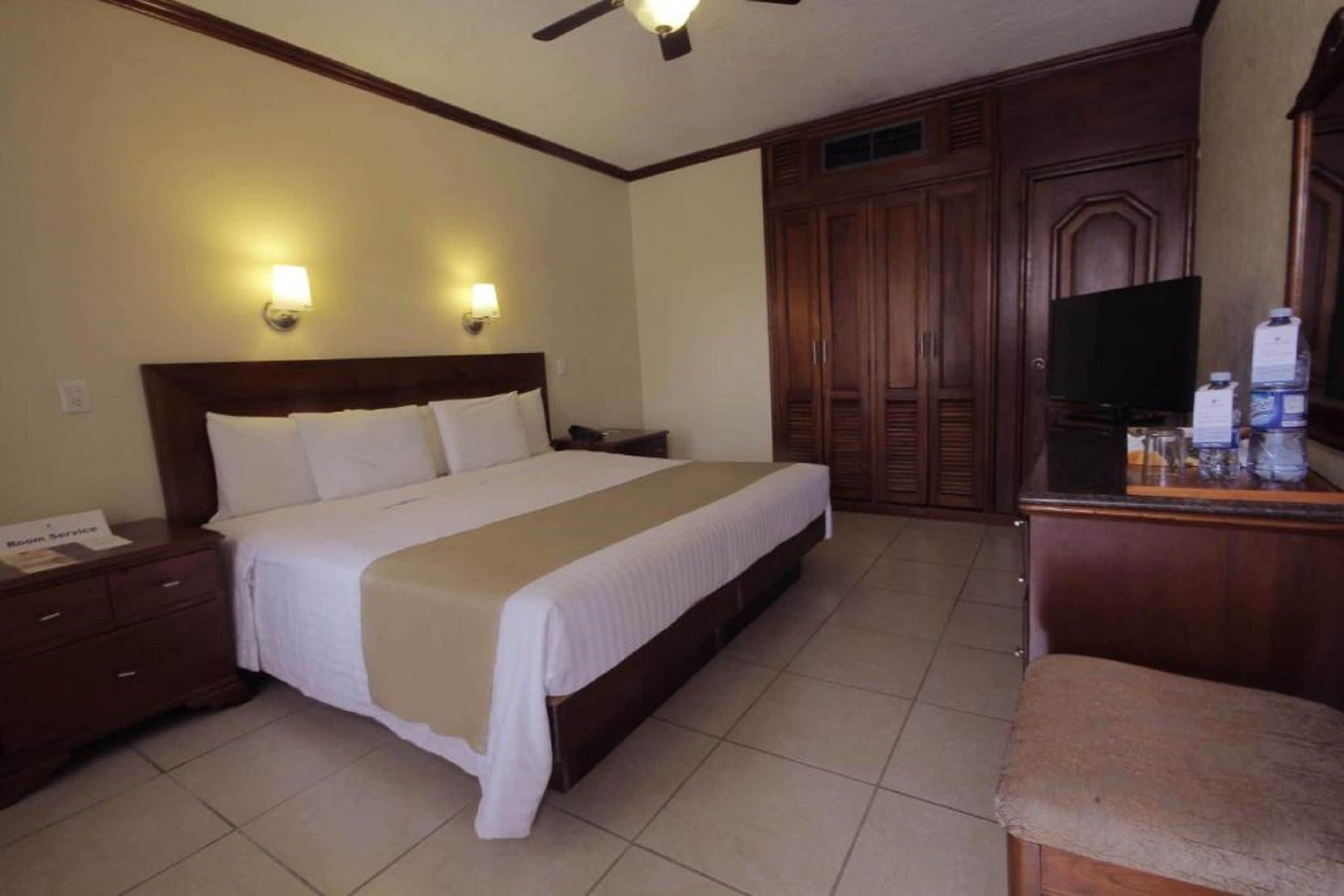 Habitación de hotel con una cama grande, una mesita de noche, un escritorio con televisor y armarios de madera.