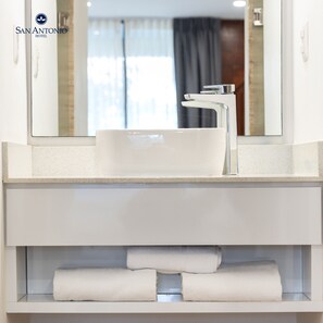 A modern bathroom with a white sink, chrome faucet, and a mirror above it. There are towels on a shelf below the sink. - Hotel San Antonio (Tampico)