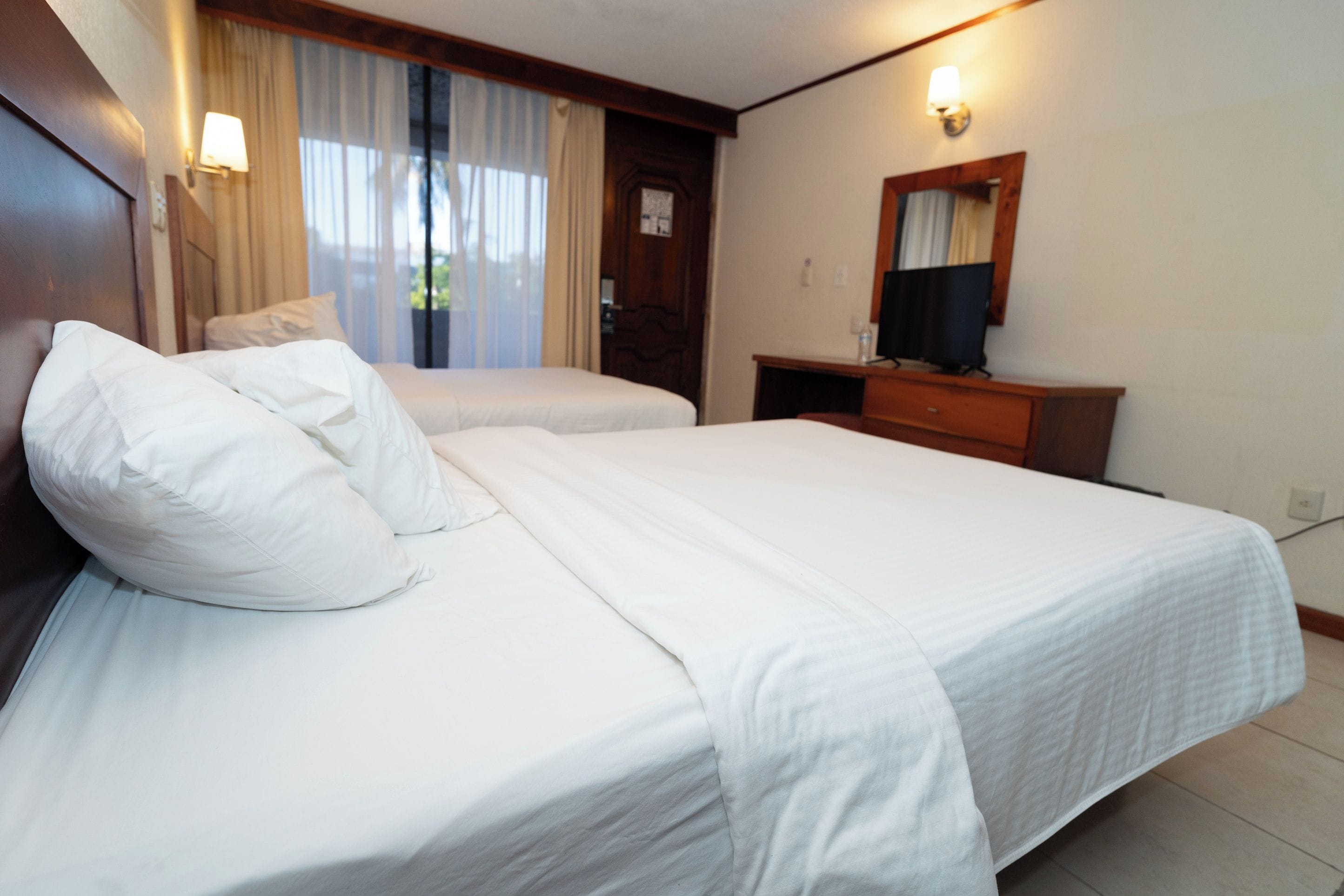 A hotel room with two beds, a desk, a television, and a window with curtains.