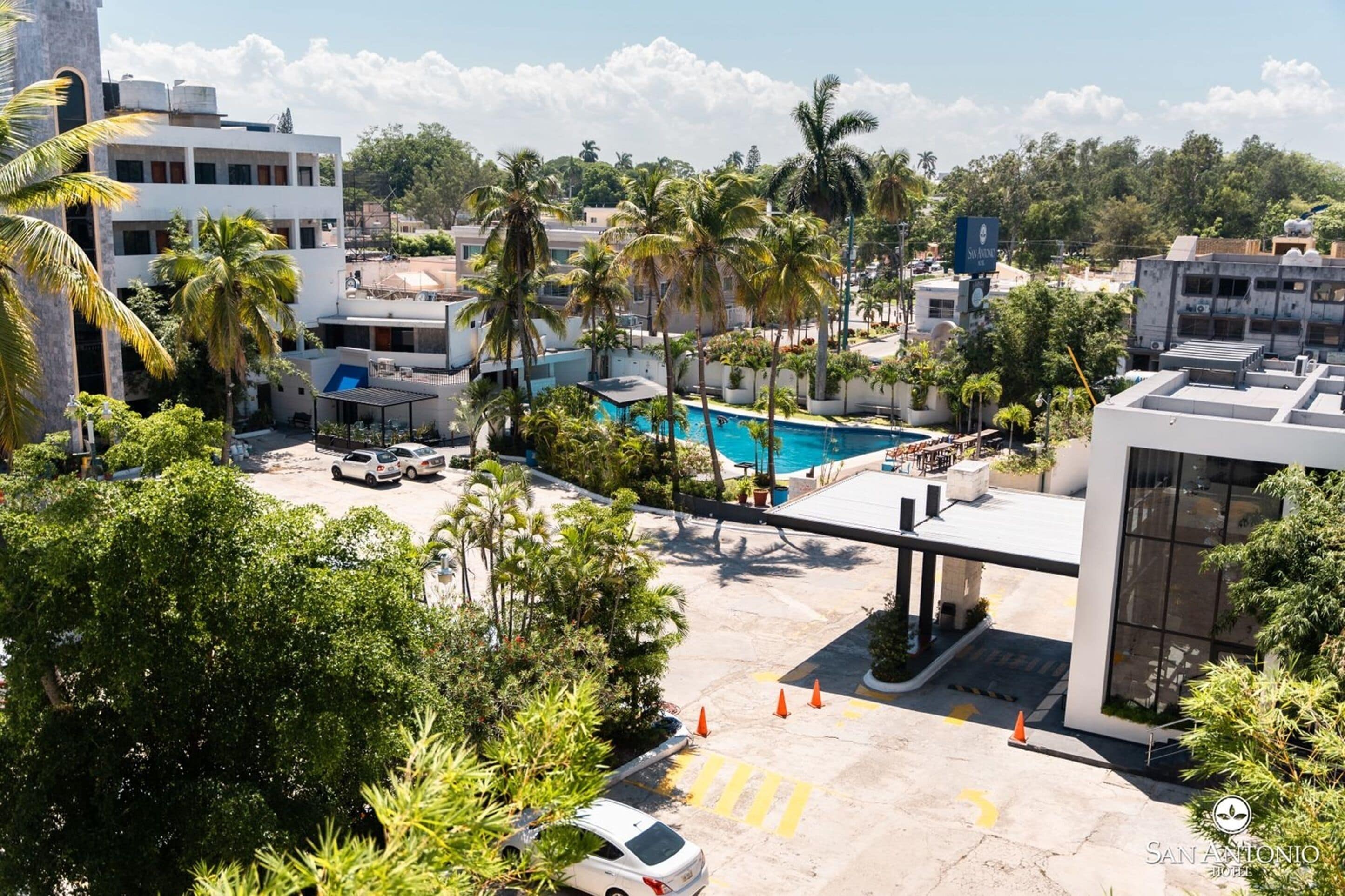 Un hotel con piscina rodeado de palmeras, zona de estacionamiento y edificios modernos al fondo.