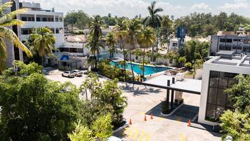 Un hotel con piscina rodeado de palmeras, zona de estacionamiento y edificios modernos al fondo.