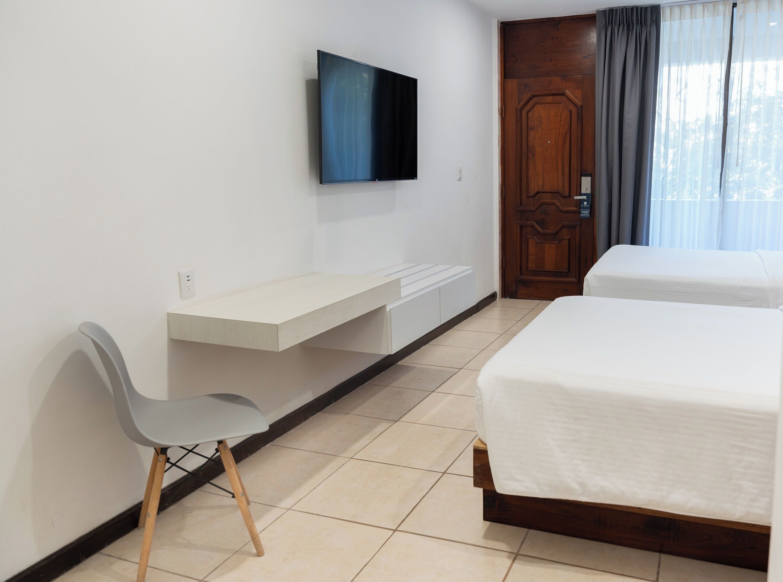 A hotel room with a bed, a desk, a chair, a TV mounted on the wall, and a wooden door.
