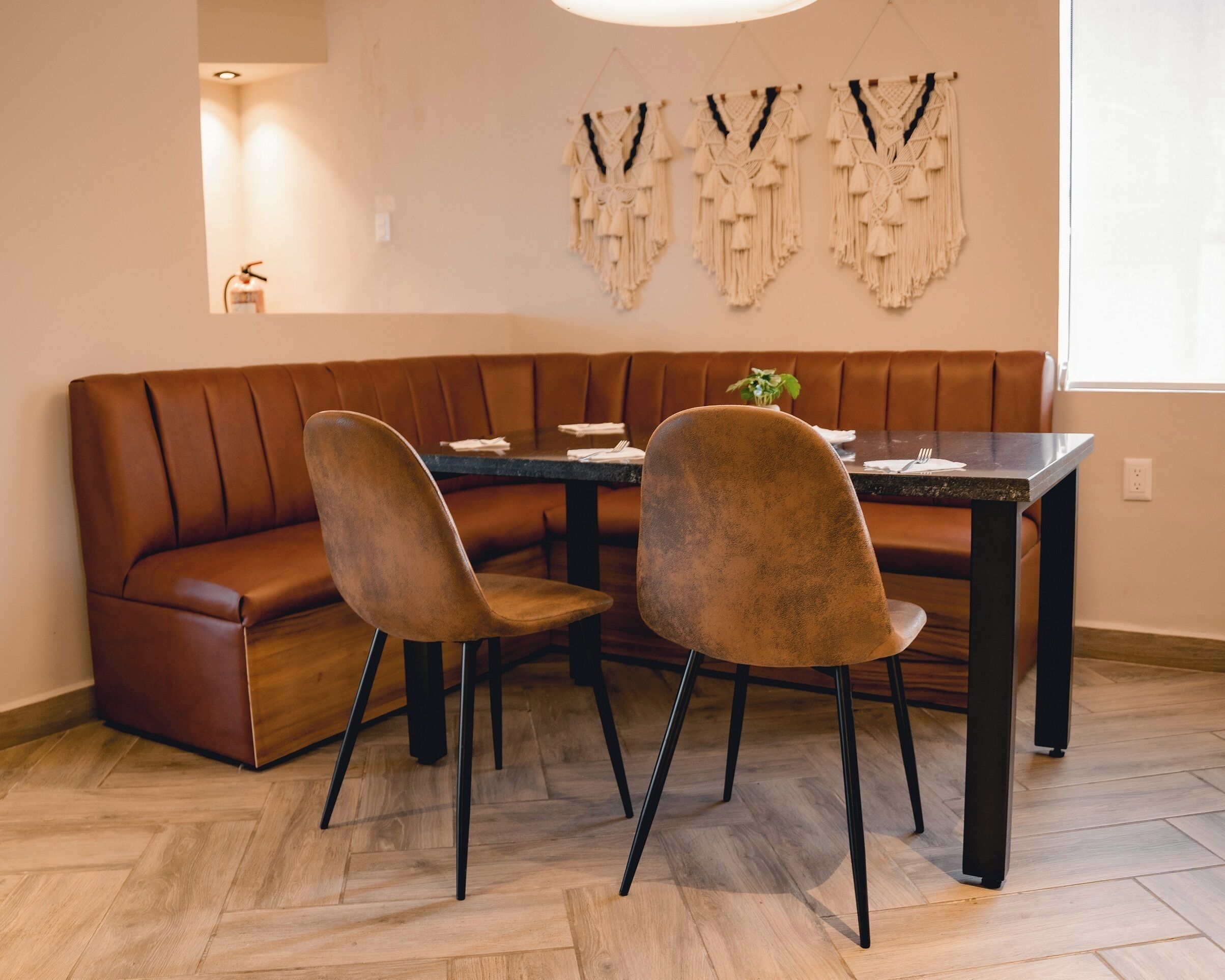 A dining area with a brown leather booth, two chairs, a dark table, and wall-mounted macramé decorations.
