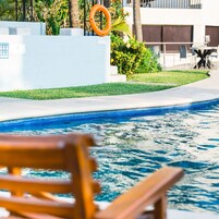 Zona de piscina con un banco de madera, un flotador salvavidas y una pared blanca con un panel de control.