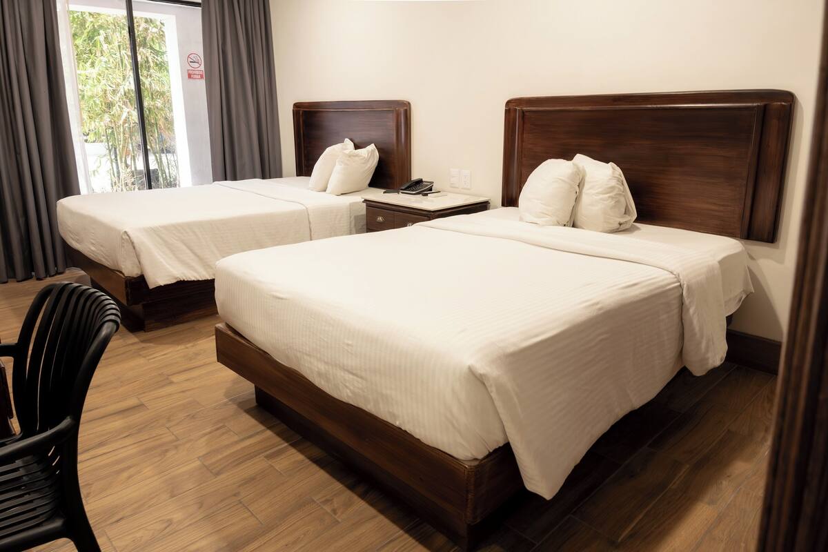 A hotel room with two beds, a wooden floor, a chair, and a window with curtains.