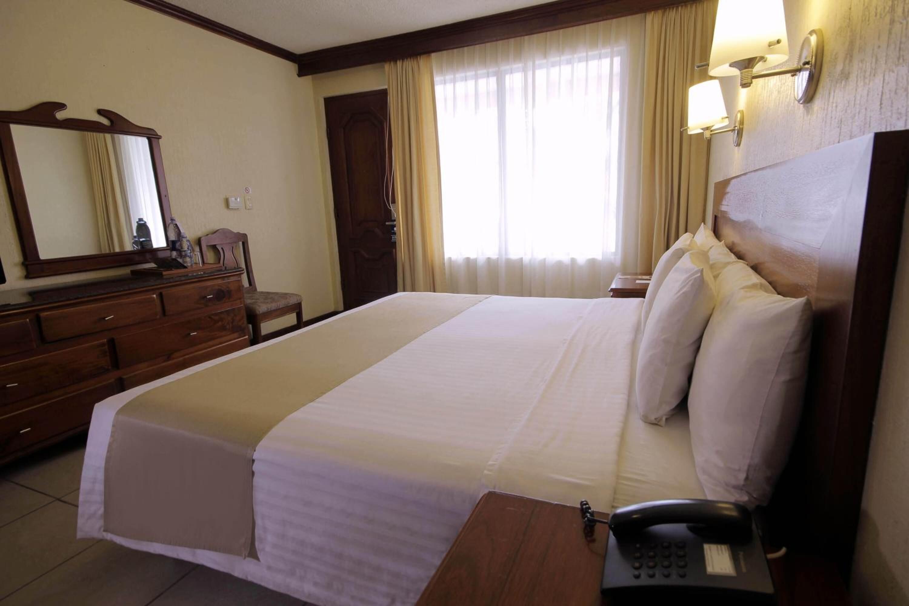Una habitación de hotel con una cama grande, un aparador de madera, un espejo, un teléfono y una ventana con cortinas.