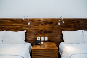 Habitación de hotel con dos camas, cabecera de madera, mesita de noche con un teléfono y un control remoto, y tres tomas de corriente.
