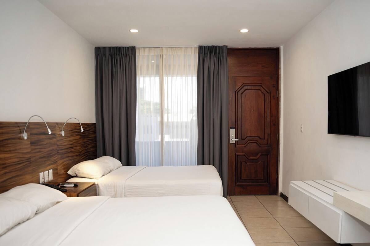A hotel room with two beds, a wooden headboard, a TV mounted on the wall, a bedside table with a lamp, and a large wooden door.