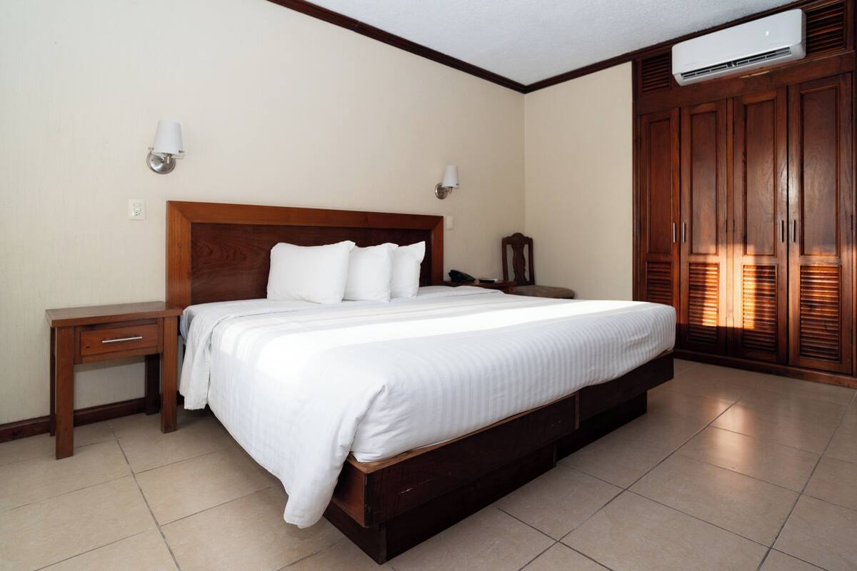 A bedroom with a large bed, wooden headboard, two bedside tables, a nightstand, a wardrobe, and an air conditioning unit.