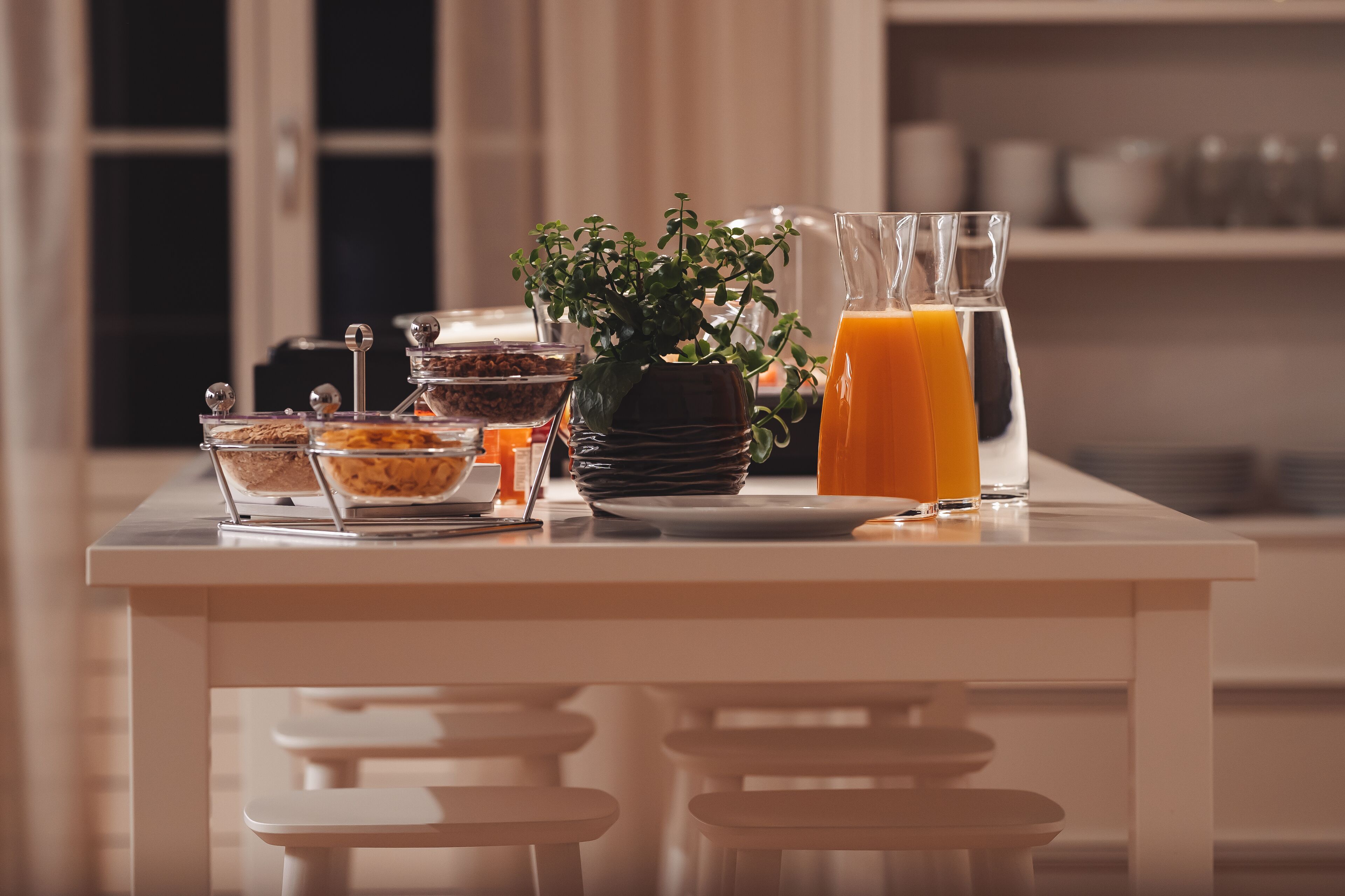 Una mesa de desayuno con una jarra de jugo de naranja, un tazón de cereal, una pequeña planta en maceta y un vaso de jugo de naranja.