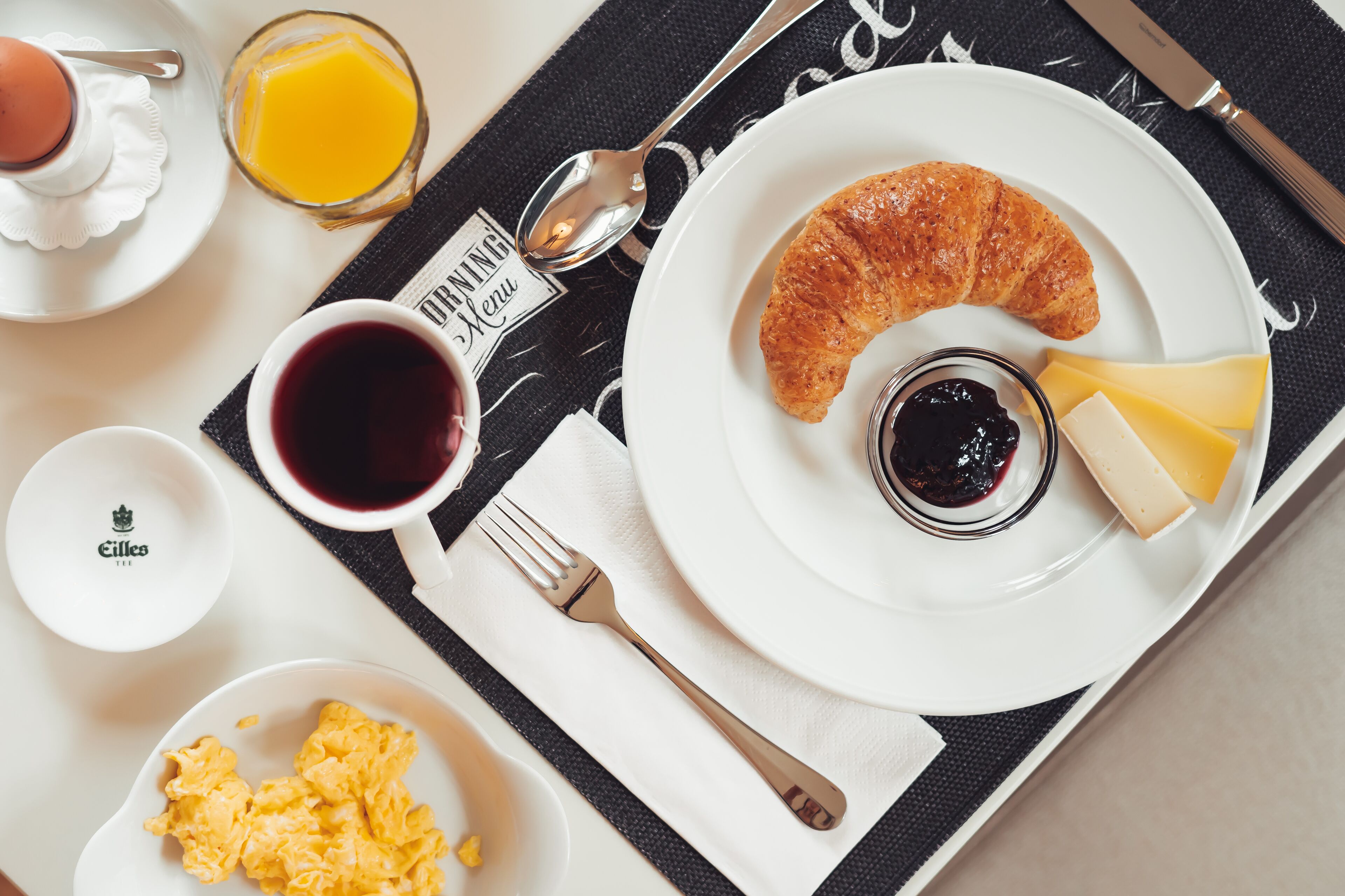 A breakfast setting with a croissant, cheese, and jam on a plate, a glass of orange juice, a cup of coffee, and scrambled eggs on a tray.