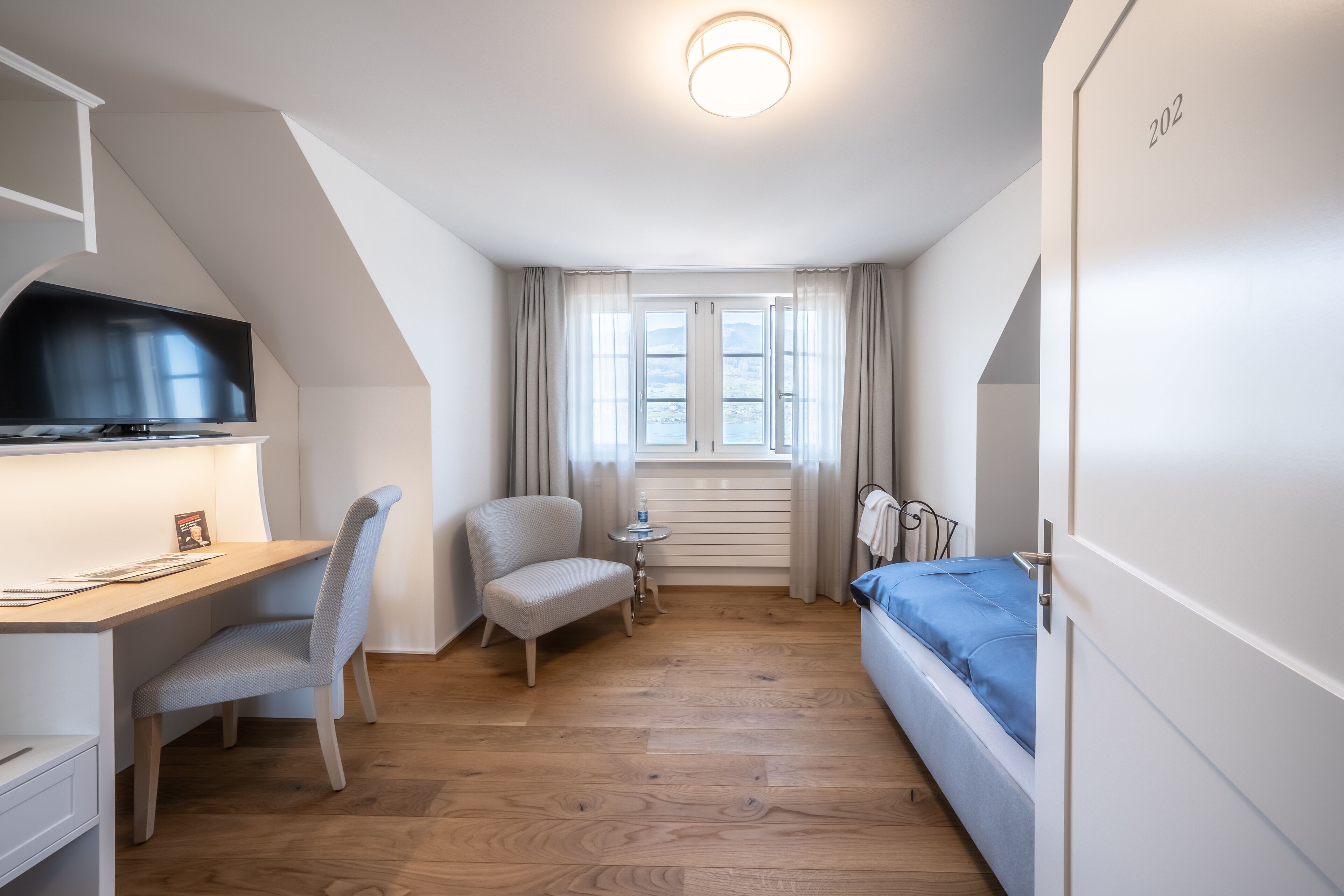 Een compacte, moderne kamer met een houten vloer, een bureau met stoel, een bed met blauwe matras, een televisie en een raam met gordijnen.